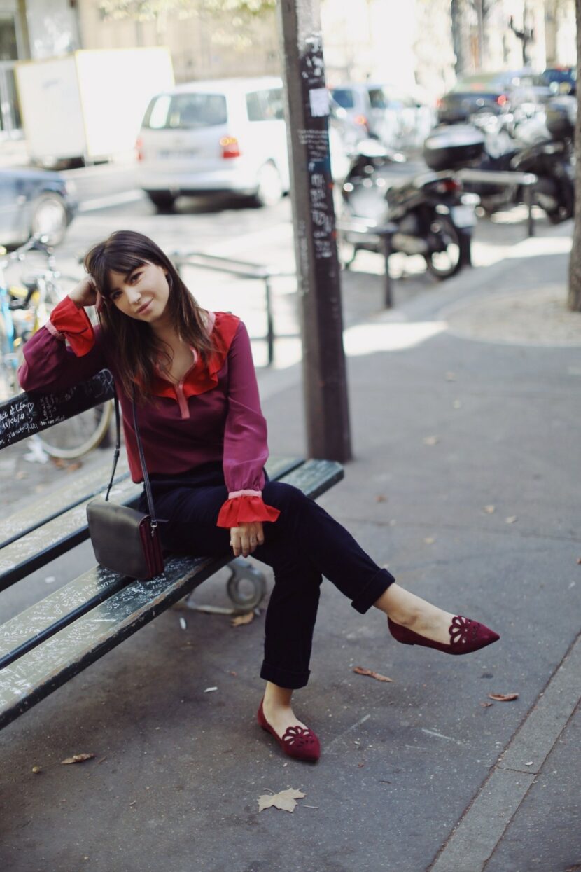 Betty Autier assise sur un banc en chemisier en soie et ballerines bordeaux