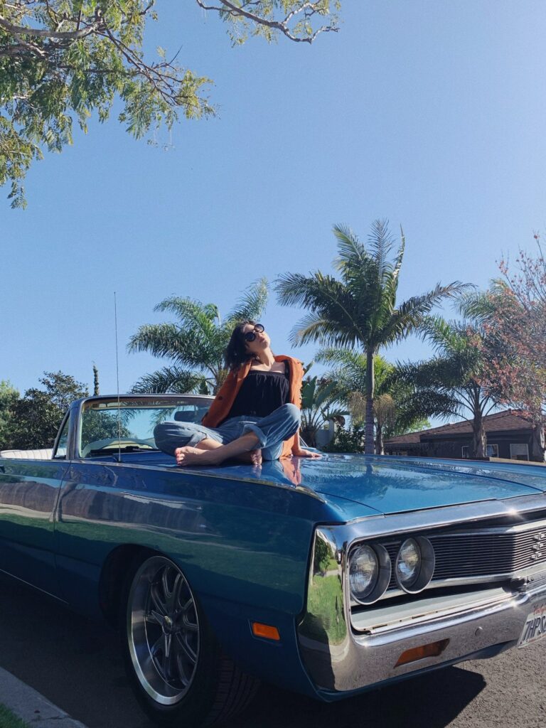 Betty Autier pieds nus sur le capot d’une voiture américaine bleue sous les palmiers de Los Angeles