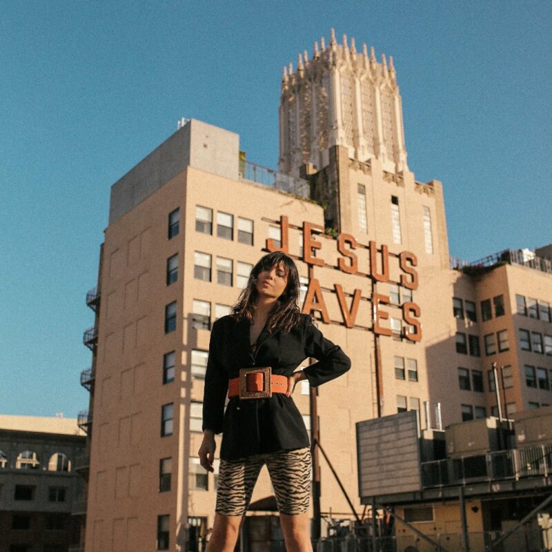 Betty Autier devant l’enseigne Jesus Saves de l’Ace Hotel à Los Angeles, blazer noir et cycliste zébré