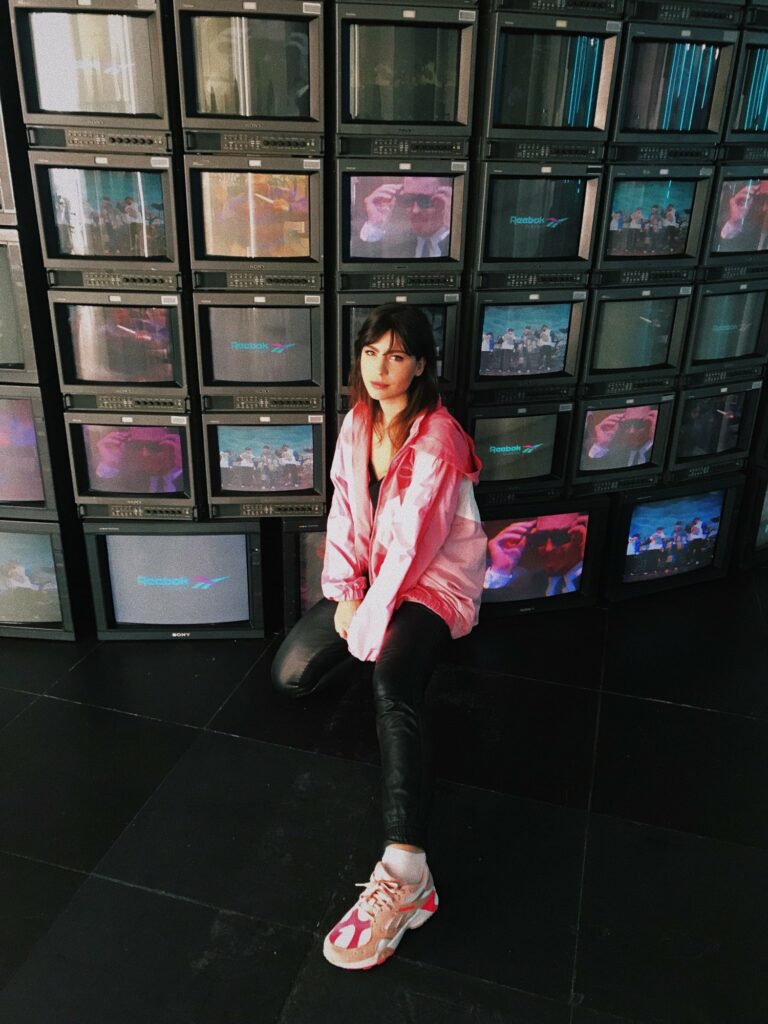 Betty Autier en coupe-vent rose devant le mur de télévisions Reebok
