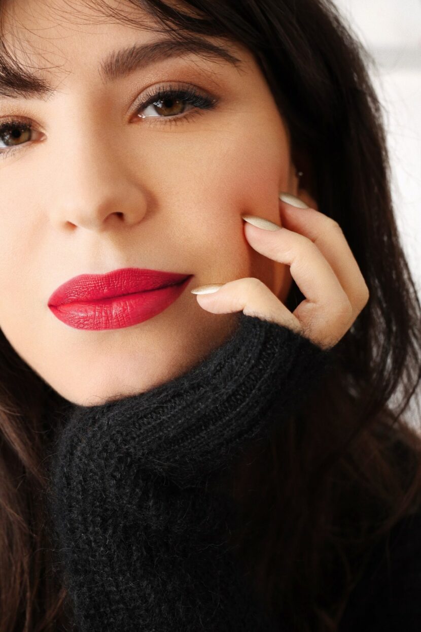 Betty Autier en pull noir, rouge à lèvres rouge et vernis doré Betty x Bourjois