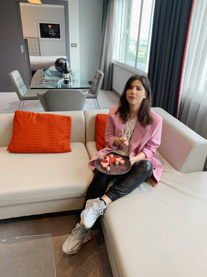 Betty Autier sur le canapé de sa suite au Pullman London St Pancras, assiette de desserts à la main