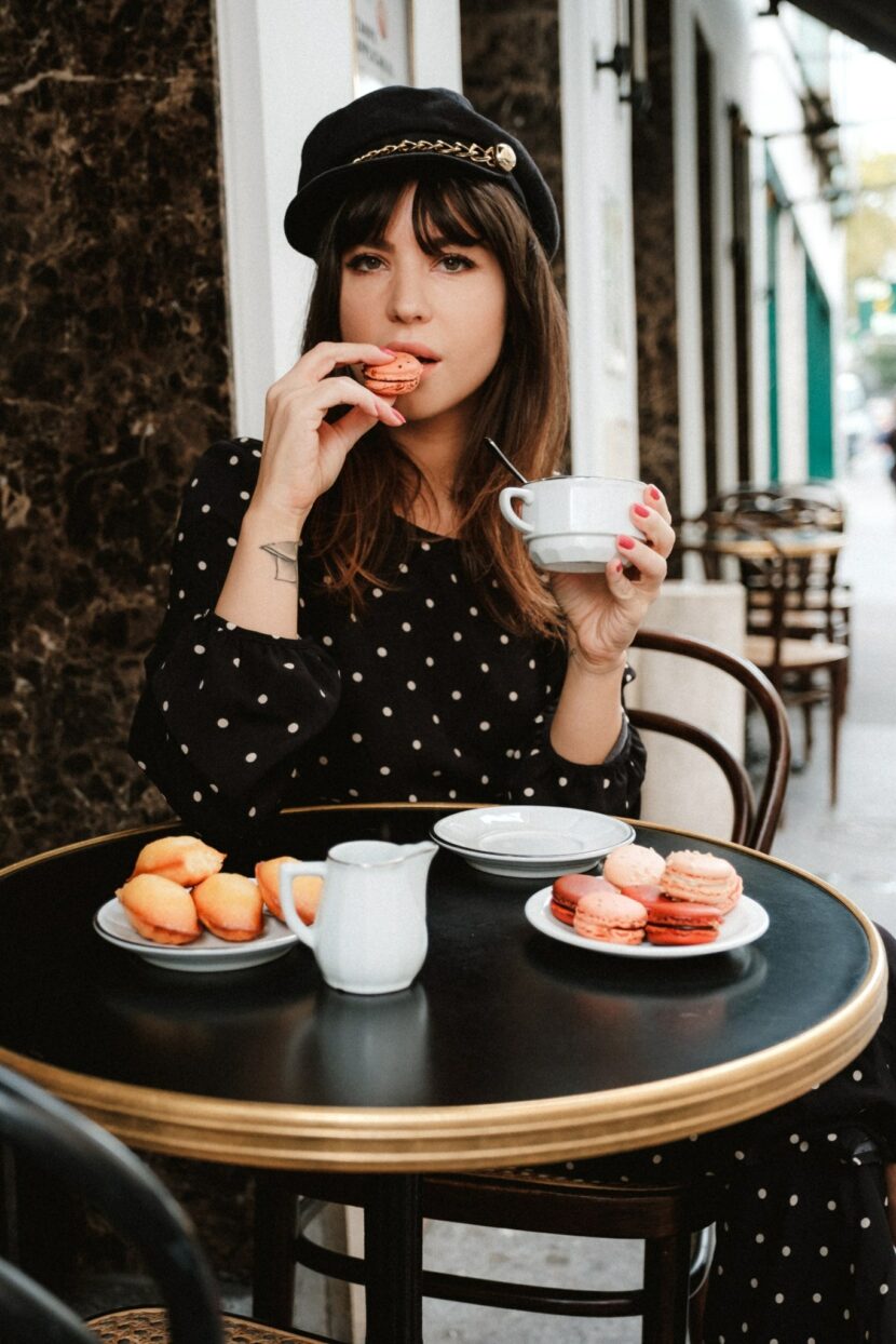Betty Autier en robe à pois et casquette, macaron à la main, en terrasse de café pour Bourjois