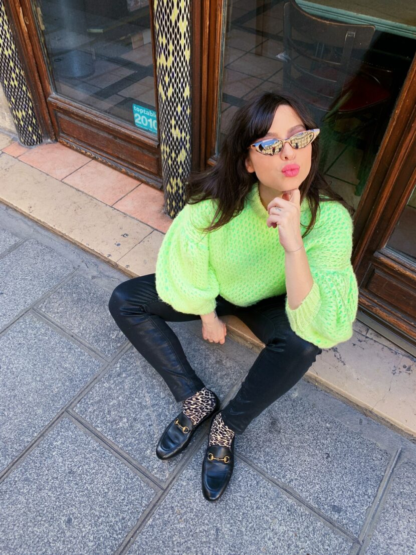 Betty Autier en pull jaune fluo, chaussettes léopard et mocassins Gucci, assise devant une porte