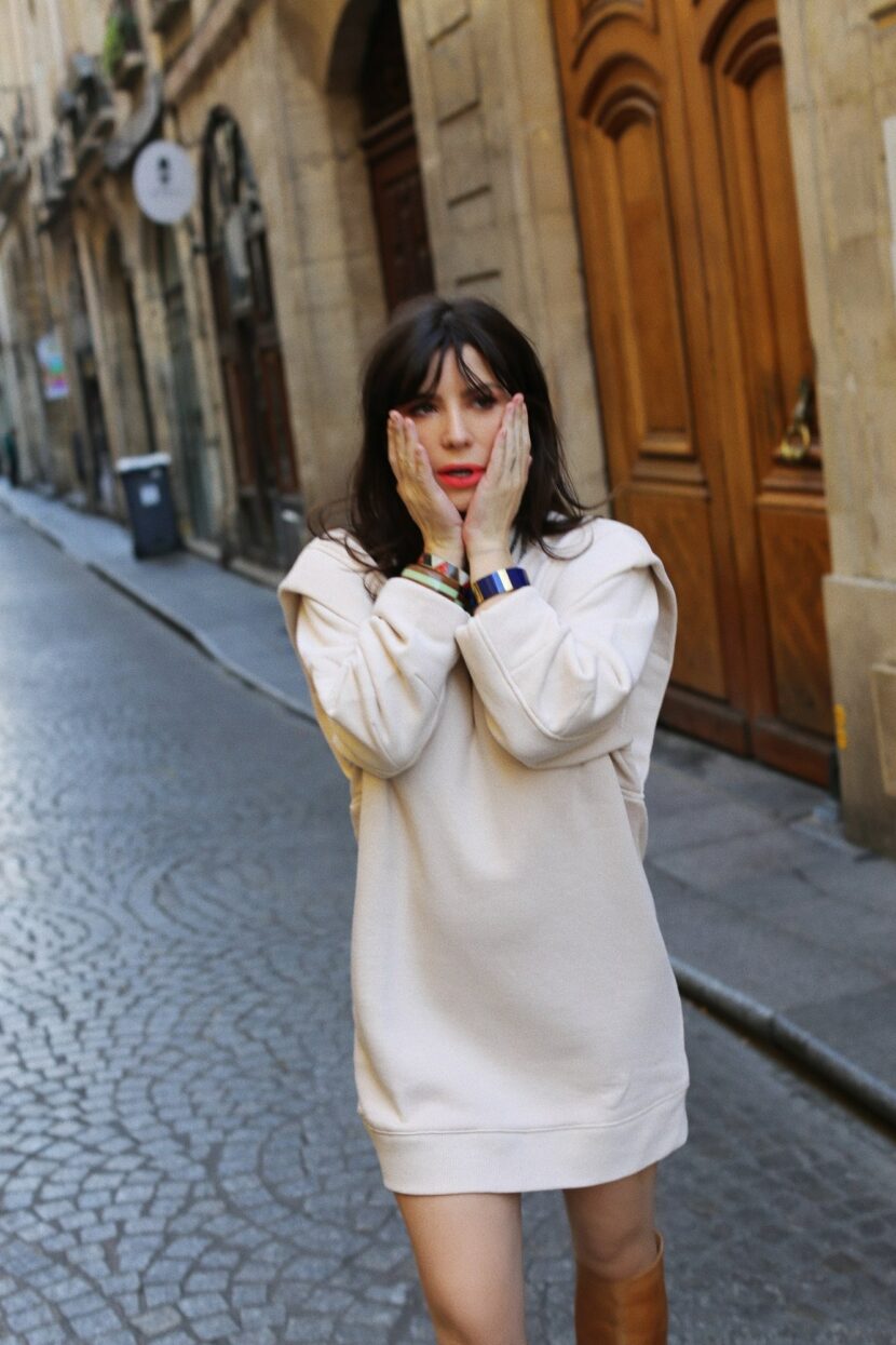 Betty Autier en robe pull écrue, mains sur les joues, dans une rue pavée de Paris