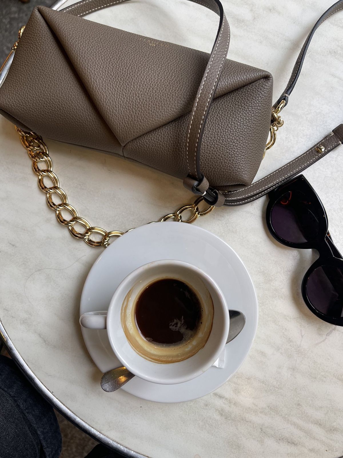 Sac Fleuron Trinity taupe avec chaîne dorée, café et lunettes de soleil en terrasse