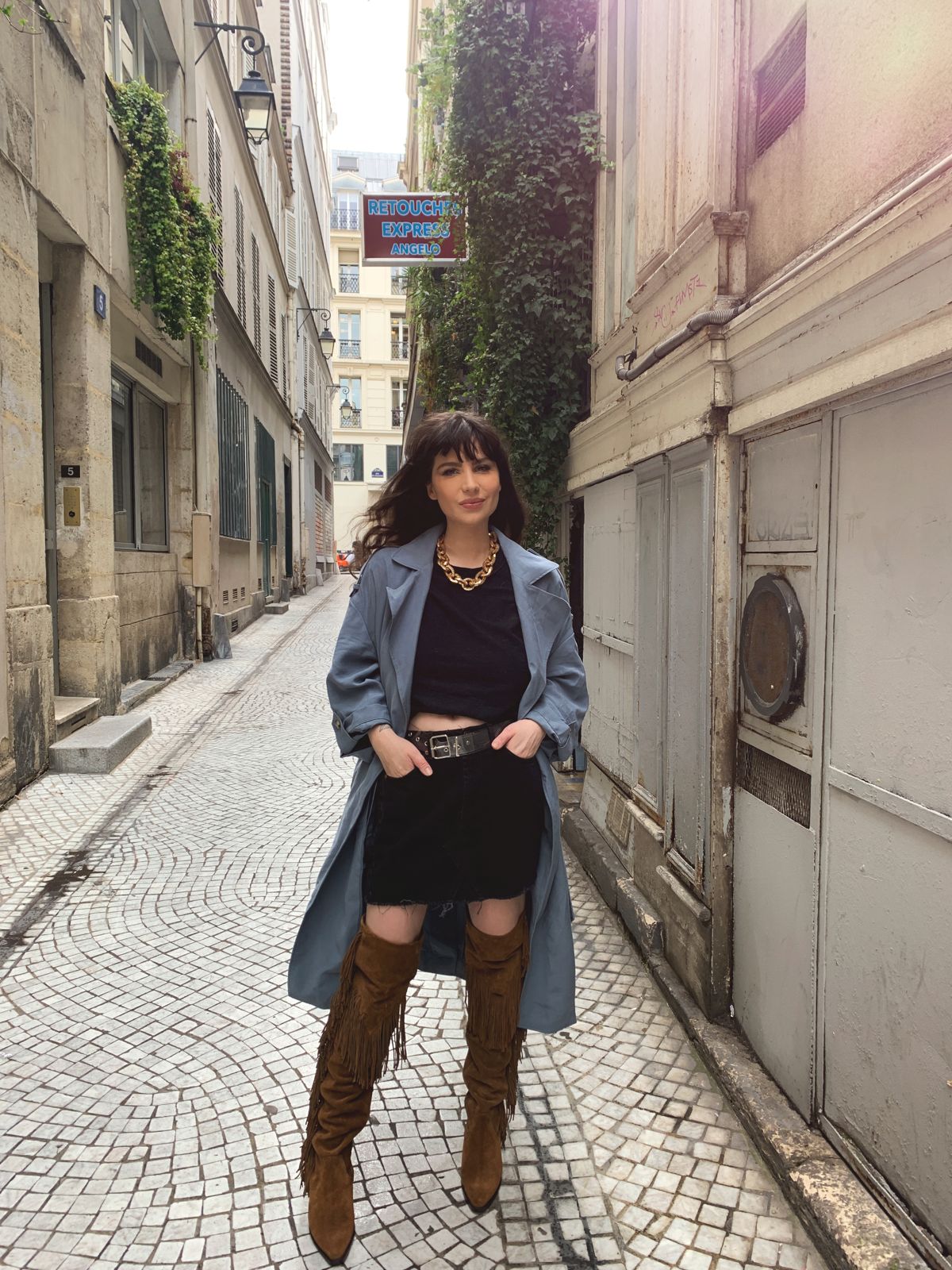Betty Autier en trench bleu, cuissardes à franges camel et collier grosses mailles dans une ruelle de Paris
