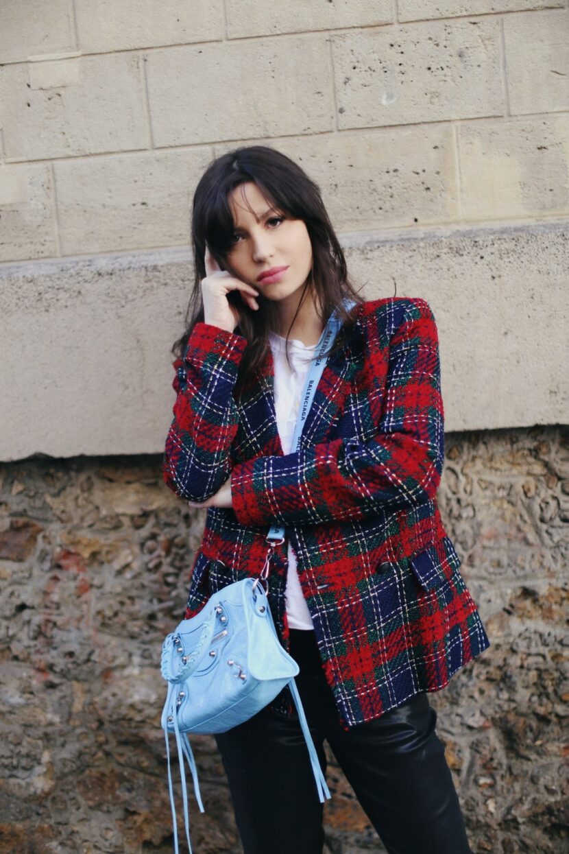 Betty Autier en veste tartan avec un sac Balenciaga City bleu, contre un mur en pierre